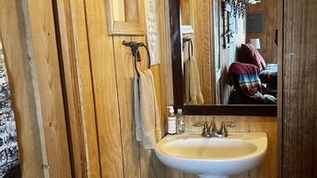 Un baño de madera con un lavamanos blanco, una toalla colgada en un perchero y un espejo que refleja una cama con una colcha roja.