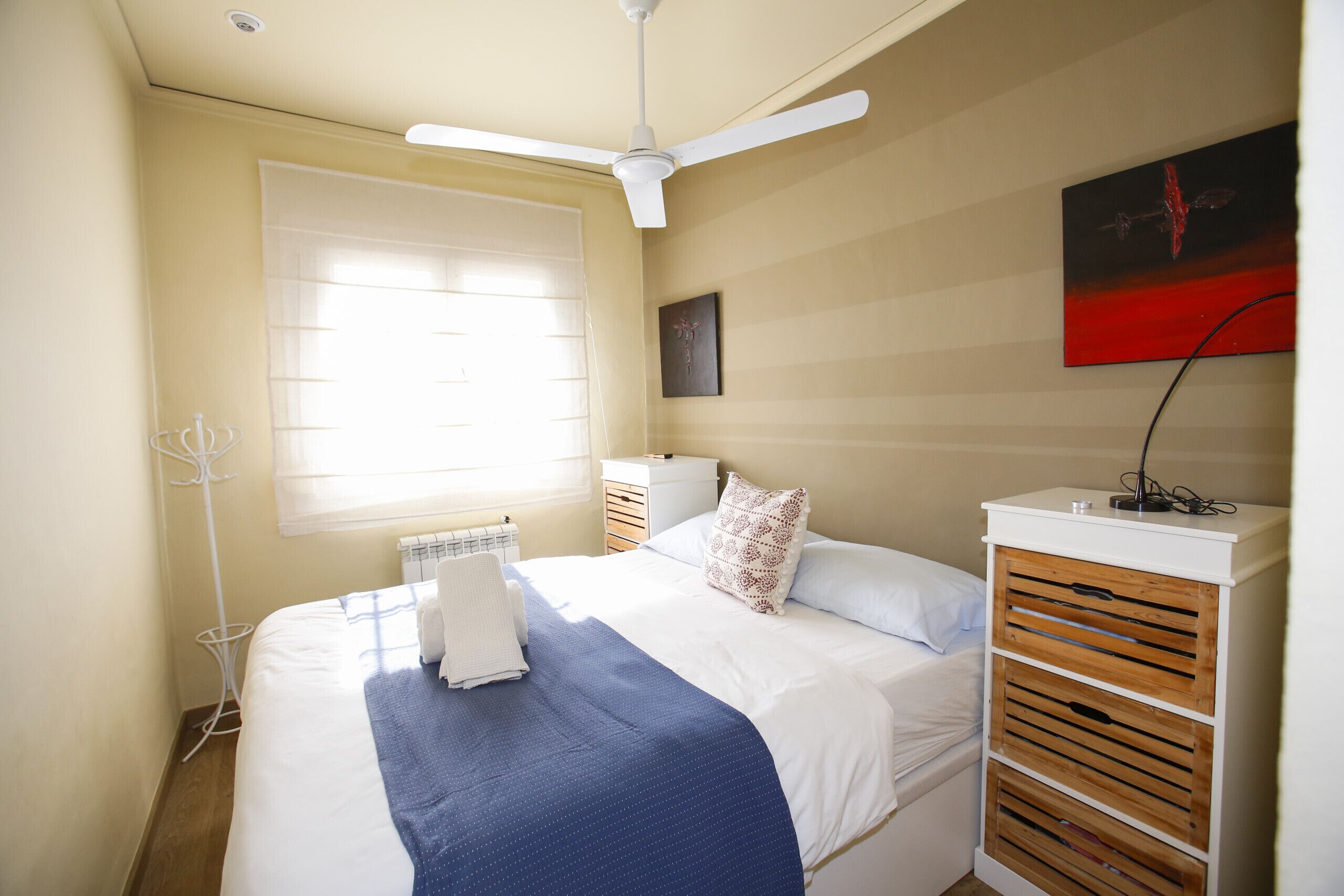 Quarto com cama, ventoinha no teto, radiador, janela e uma obra de arte na parede.