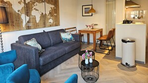 Ein modernes Wohnzimmer mit grauem Sofa, blauen Sessel, einem Holztisch mit Vasen, einem Essbereich mit Holztisch und Stühlen sowie einer Küchenzeile mit Herd und Spüle.