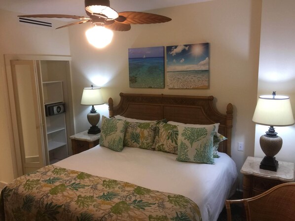 Habitación de hotel con cama, dos lámparas de noche, ventilador de techo, mesita de noche con radio y dos cuadros en la pared de una playa y un atardecer.