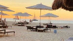 A beach with lounge chairs and umbrellas, a sailboat in the background, and a thatched roof structure. - ON EAGLE BEACH Costa Linda Beach Resort (oranjestad)