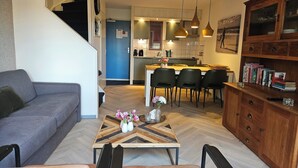 A modern living room with a sofa, a coffee table with flowers, a dining area with chairs, and a kitchen in the background.
