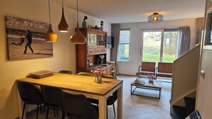 A dining area with a wooden table and chairs, a kitchen area with cabinets, a dining table with a vase of flowers, and a view of the outdoors through a window.