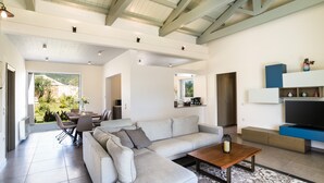 A modern living room with a sectional sofa, a coffee table, and a TV. There is a dining area with chairs and a table, and a kitchen area with cabinets and a countertop. - Three Stars Luxury Villas (Corfu)