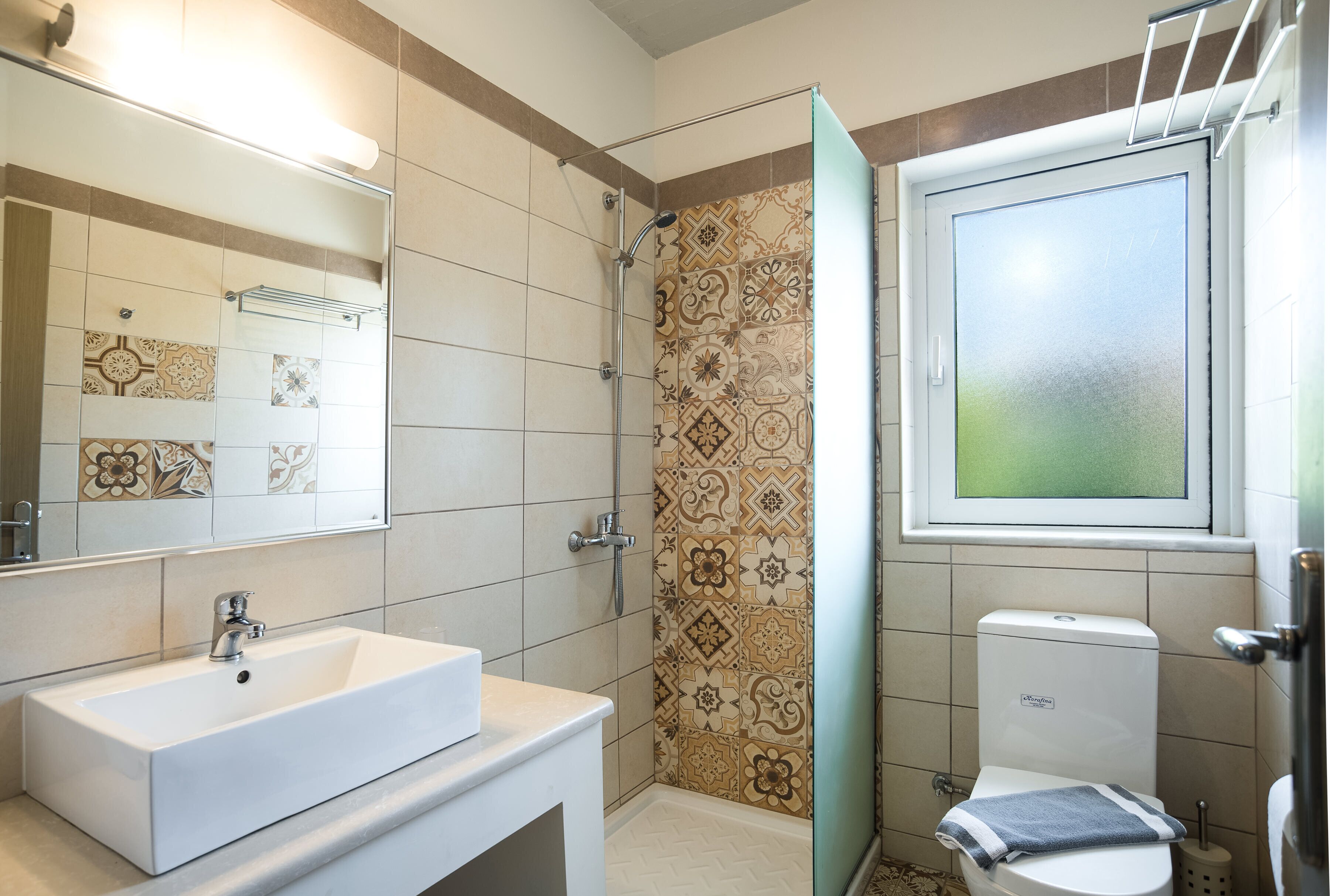 Un baño con un lavamanos blanco, una ducha con azulejos estampados y un inodoro con un ventanal encima.