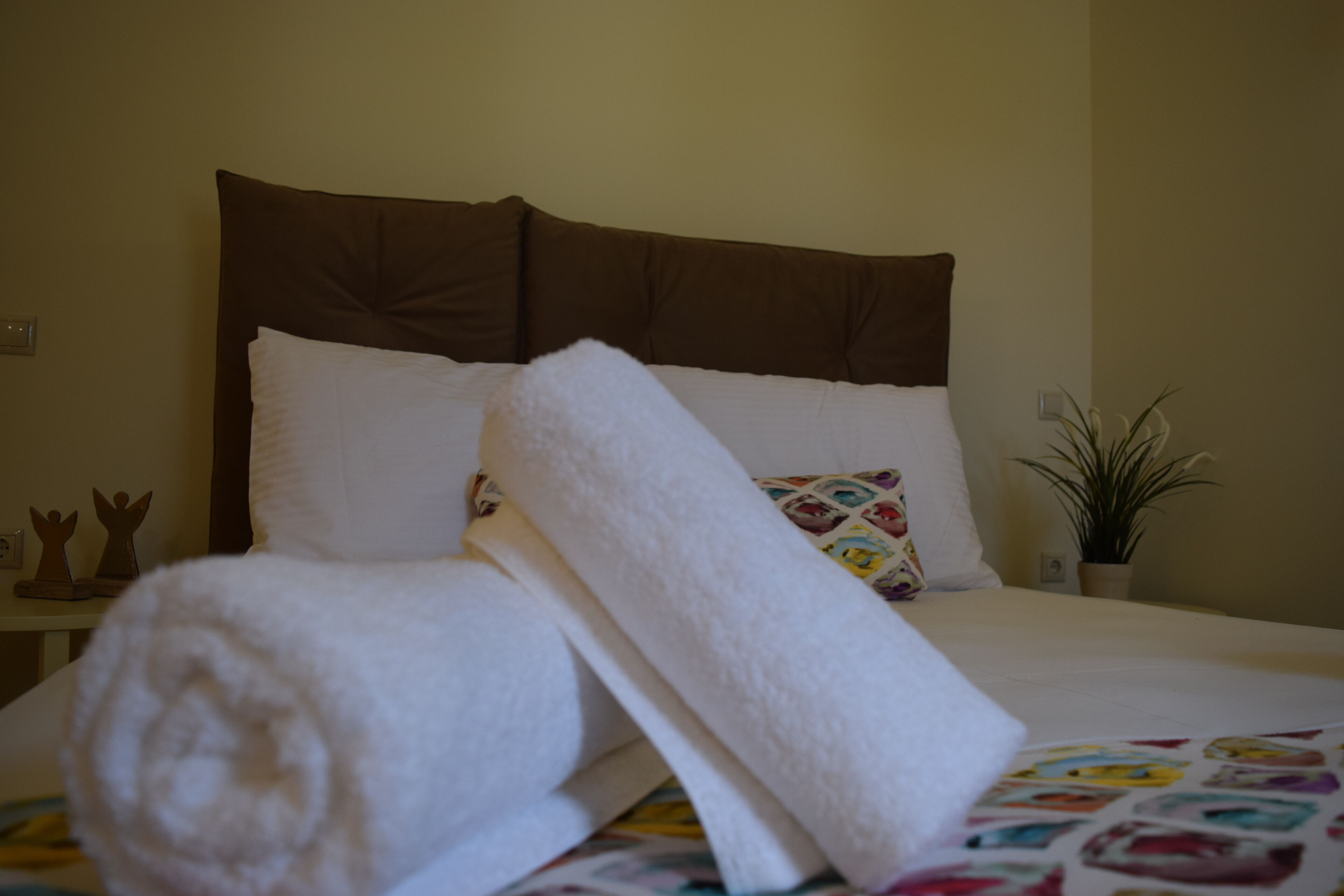 A neatly made bed with white linens and a rolled towel, a decorative pillow, and a small plant on the nightstand.