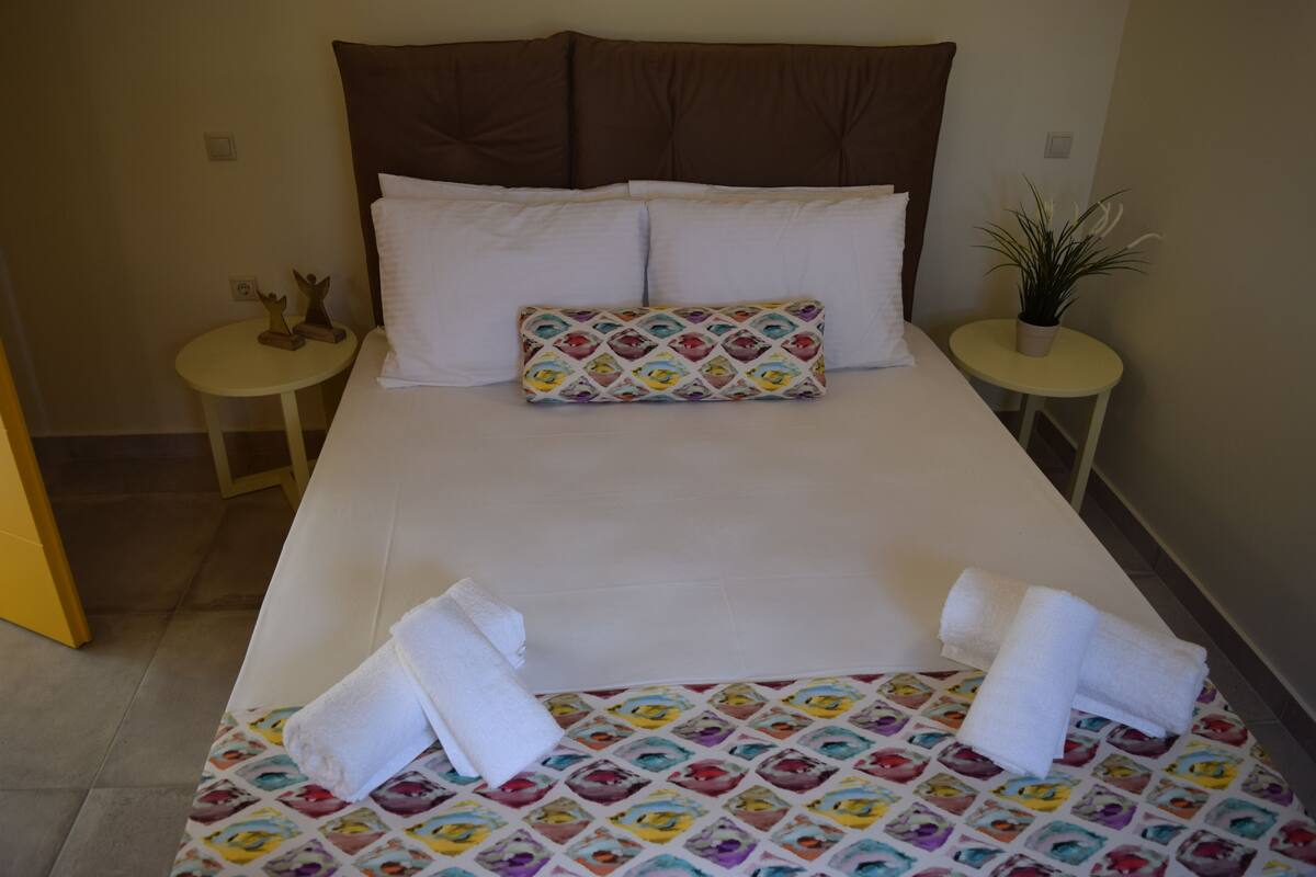 Una cama bien hecha con sábanas blancas, una almohada colorida y dos mesitas redondas con adornos.