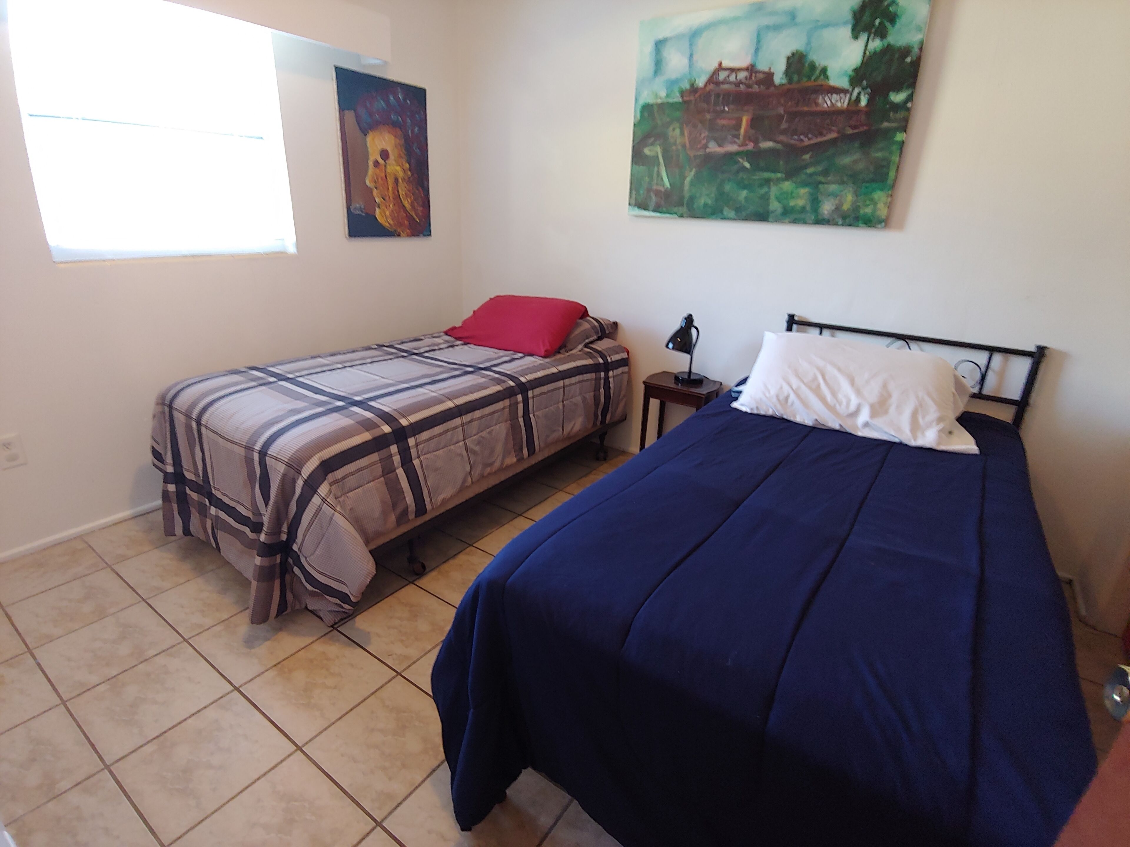 Un dormitorio con dos camas, una con cubrecama de cuadros y la otra con cubrecama azul liso. Se ve una ventana al lado izquierdo, un cuadro en la pared y una lámpara en la mesita de noche.