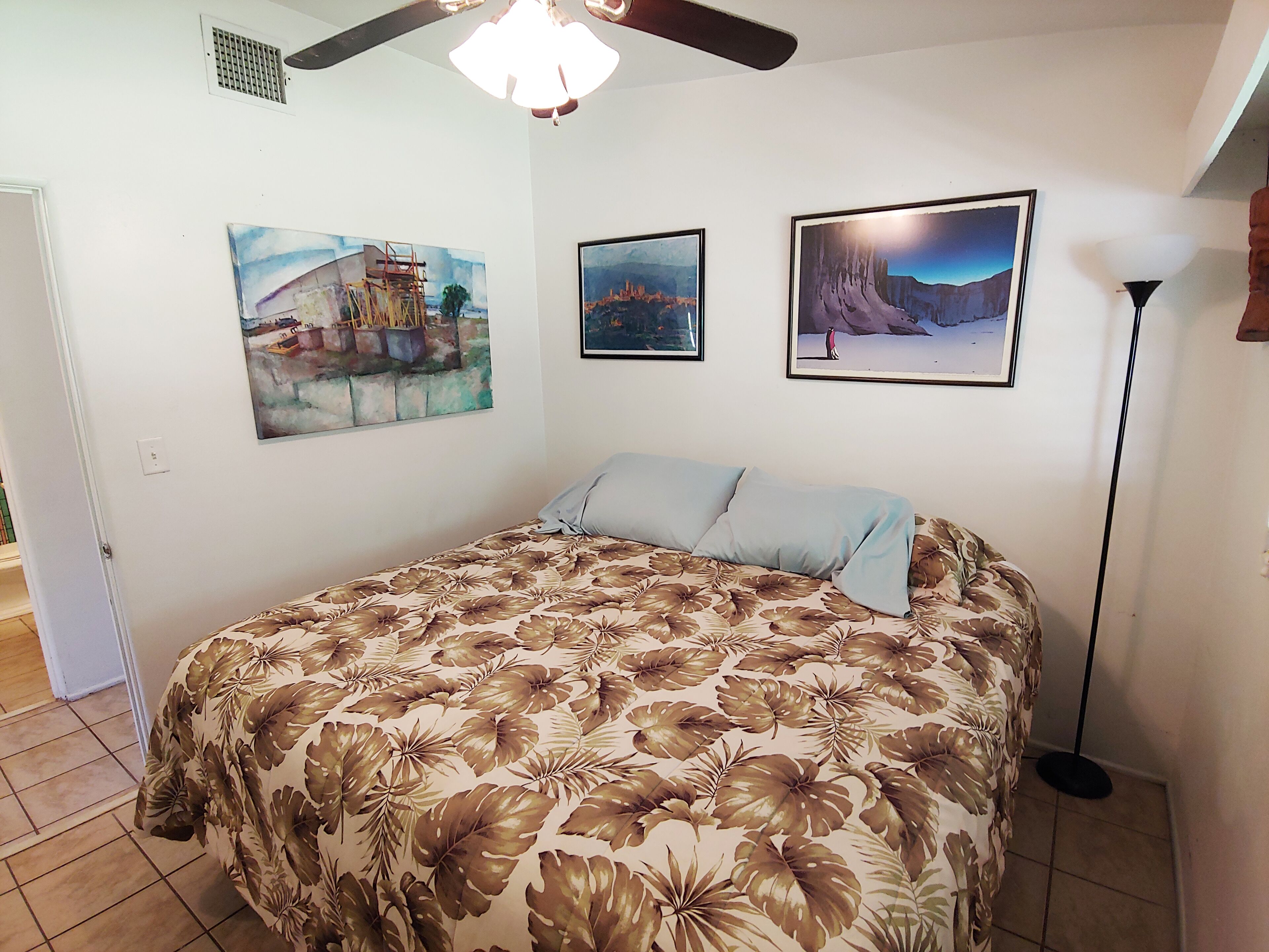 Ein Schlafzimmer mit einem blumengemusterten Bettüberwurf, einem Deckenventilator und drei gerahmten Landschaftsbildern an der Wand.
