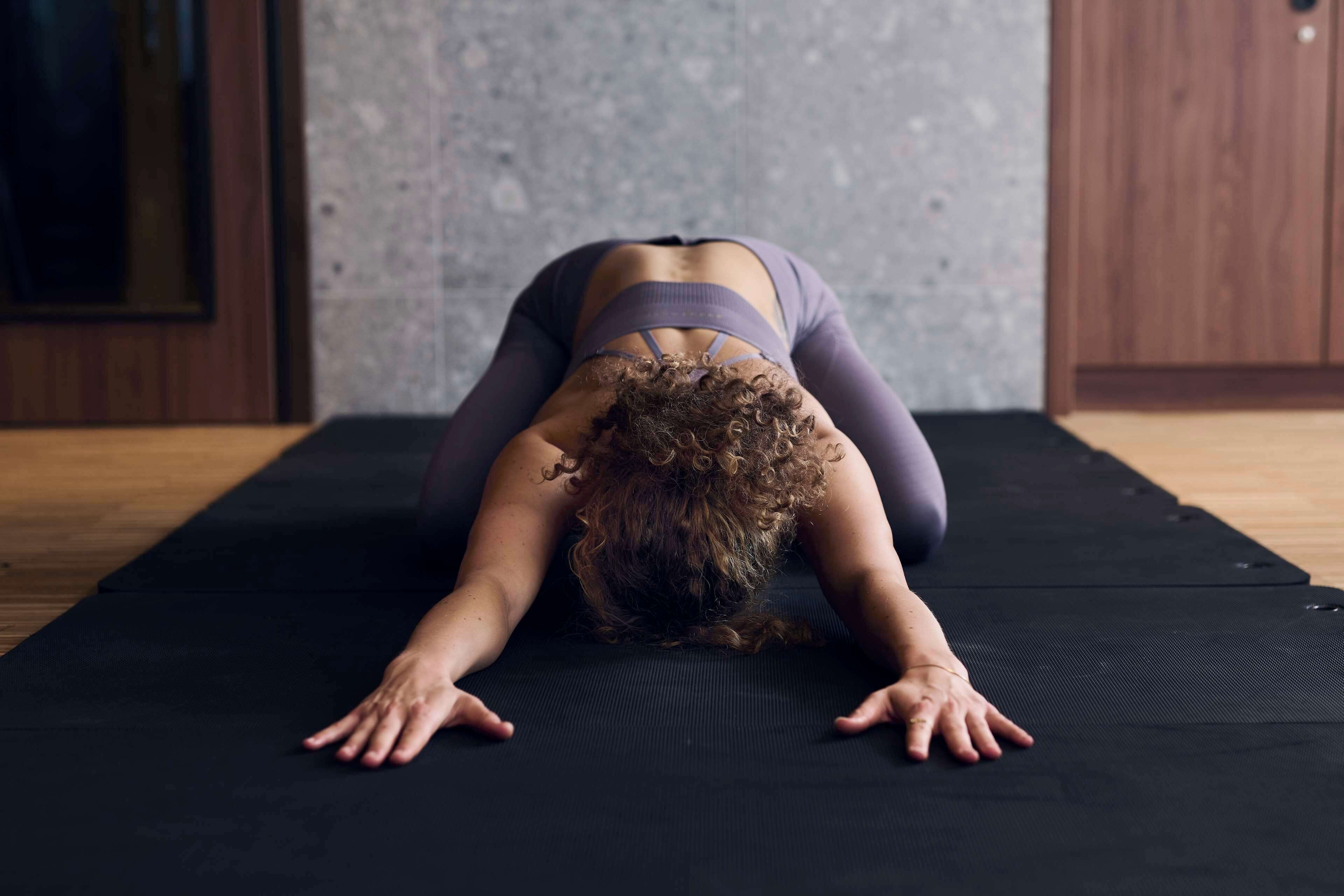 A person in a yoga pose on a black mat in a room with a wooden floor and a gray wall.