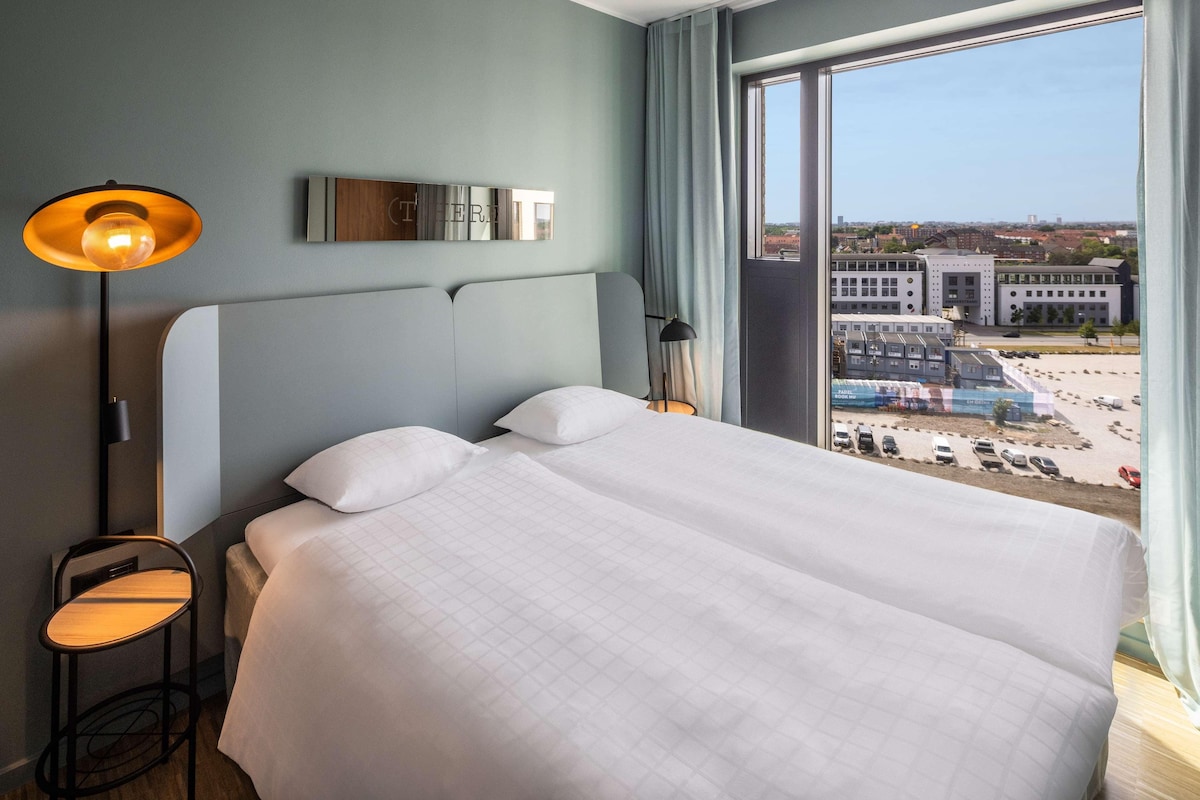A modern hotel room with a large bed, a bedside table, a lamp, and a view of the city through a large window.