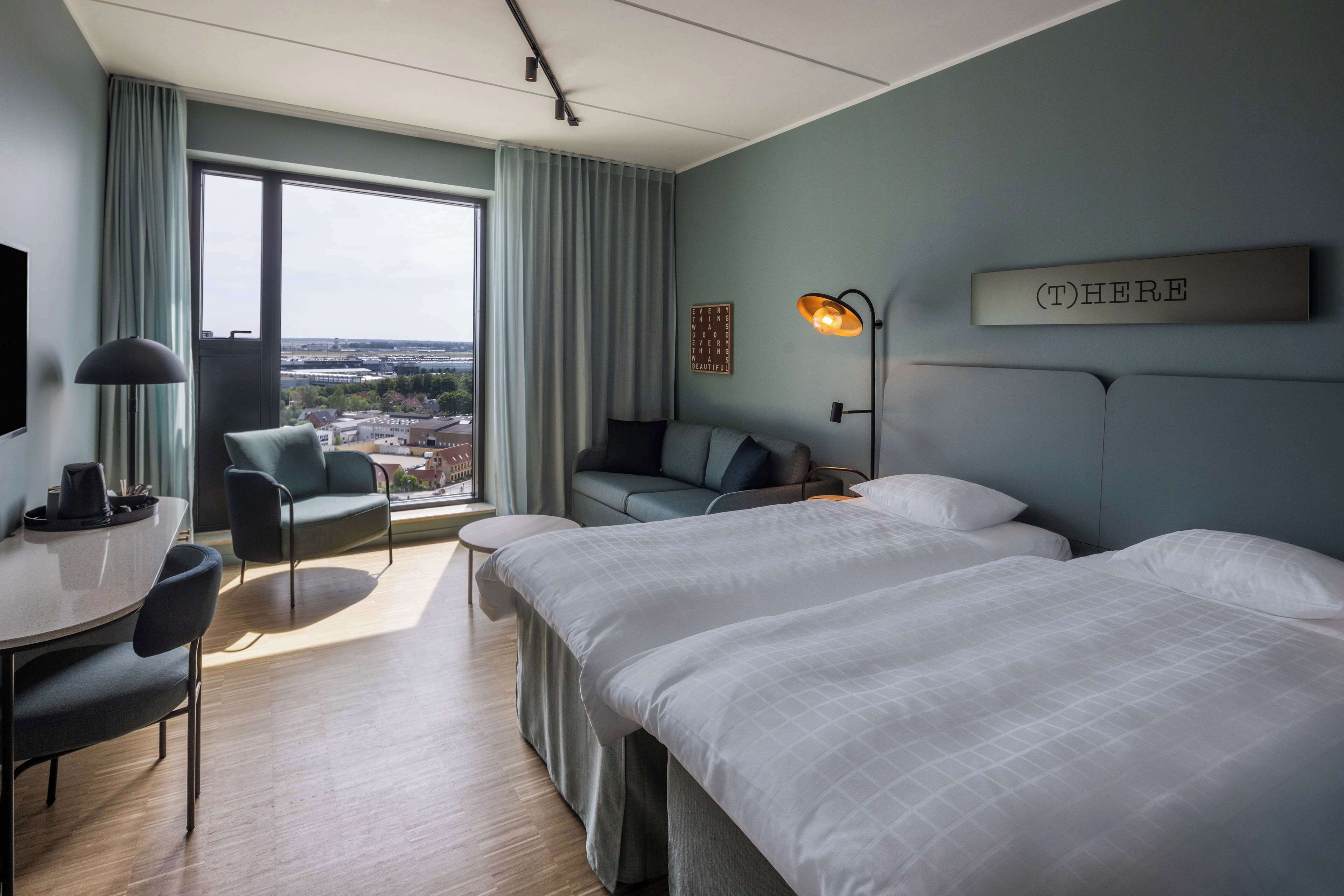 A hotel room with two beds, a desk, a chair, a sofa, a large window with curtains, a city view, a wall-mounted lamp, and a sign that says "HERE".