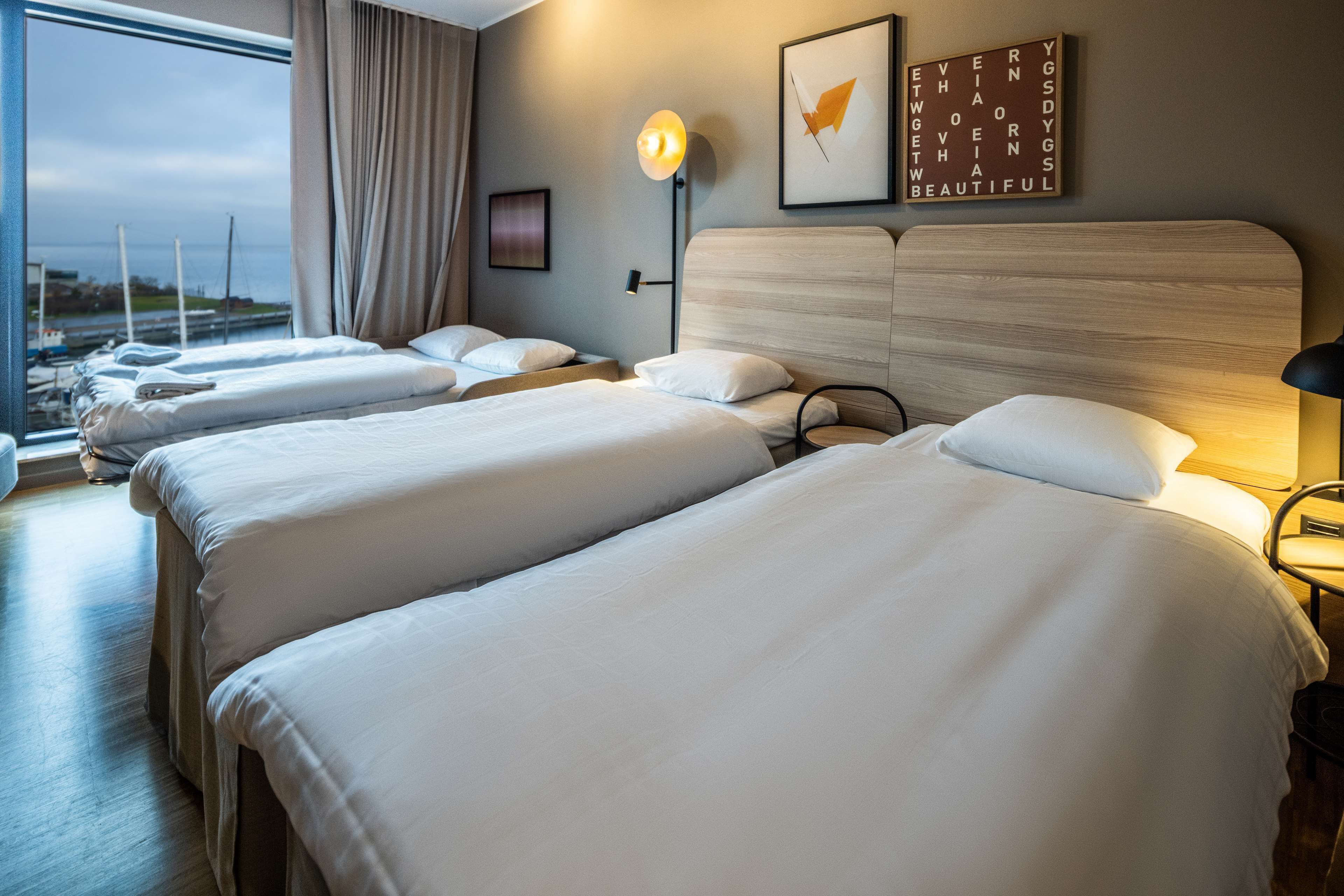 A hotel room with three beds, a large window with a view of the sea, a wall-mounted light, and a framed inspirational quote.