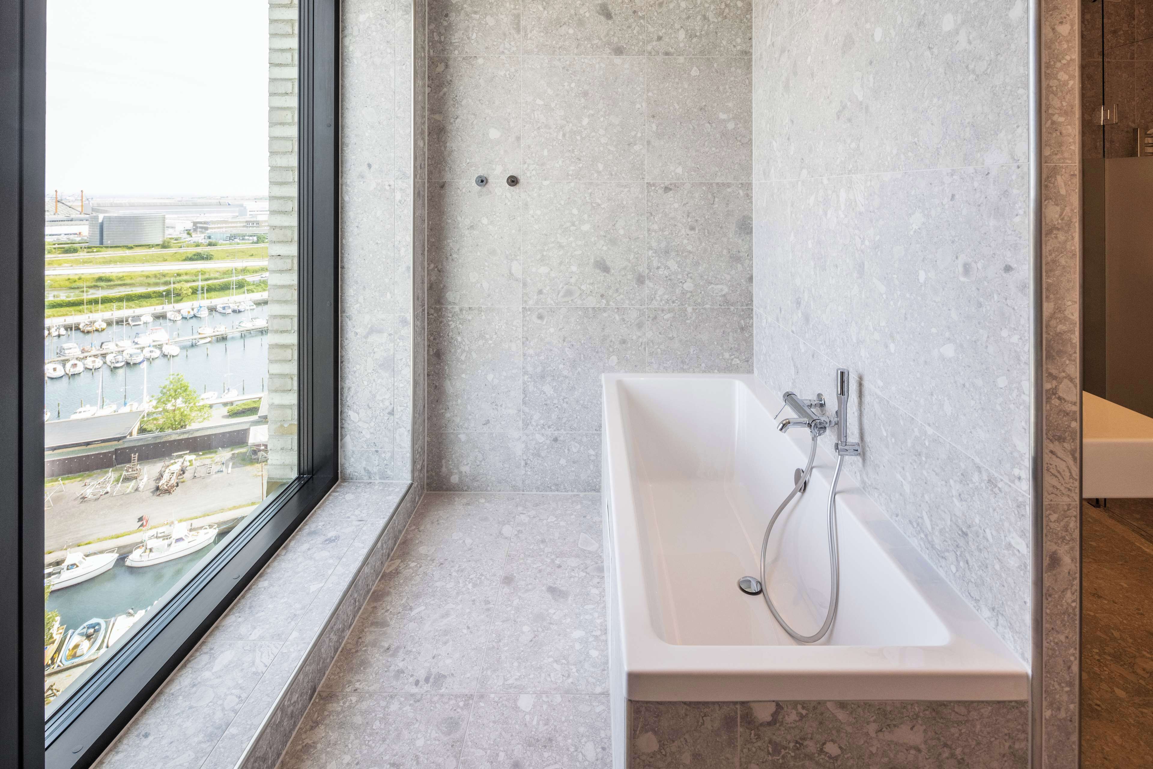 Een moderne badkamer met een groot bad, een raam met uitzicht op boten en een jachthaven, en een betegelde vloer.