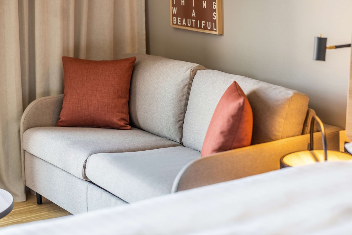 A modern living room with a grey sofa, two red cushions, a wooden side table, and a wall-mounted lamp.
