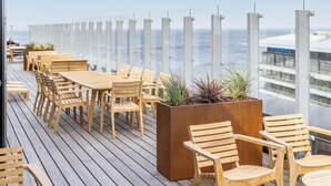 A wooden deck with tables and chairs, a planter with plants, and a view of the ocean. - Scandic CPH Strandpark (Kastrup)