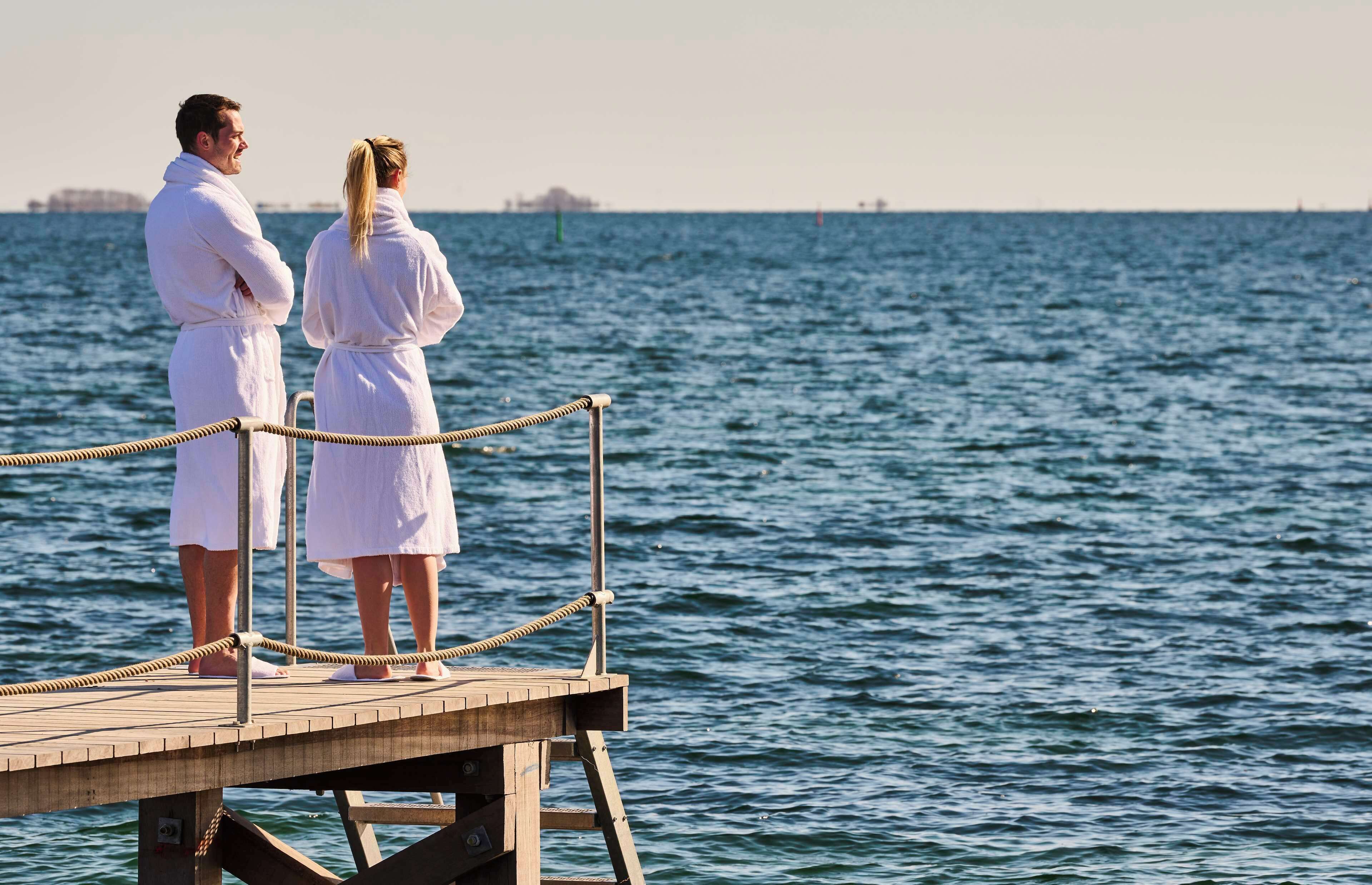 Dos personas vestidas de blanco de pie sobre un muelle de madera, mirando al mar.