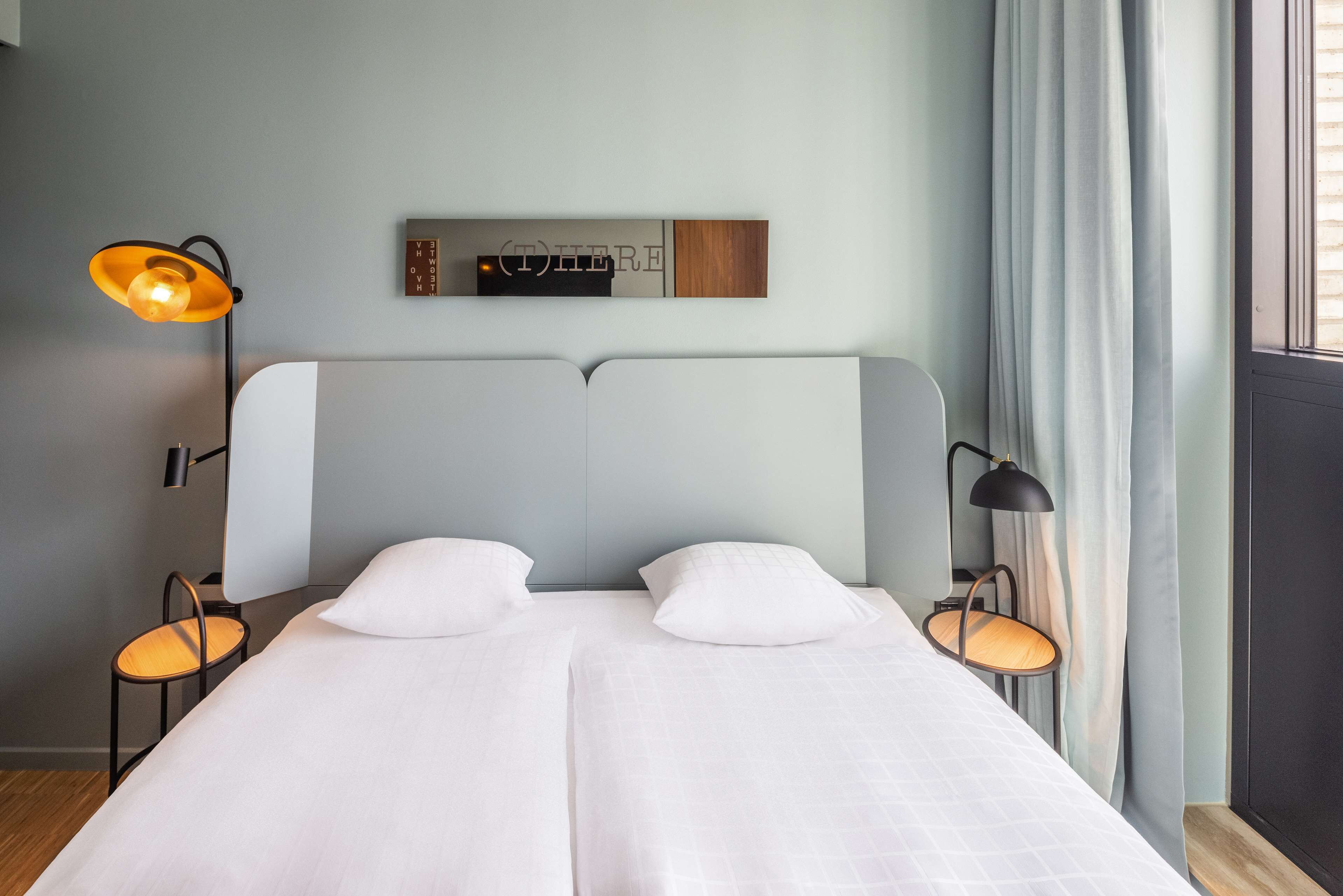 Un lit double avec du linge de lit blanc, deux tables de chevet avec des lampes, une étagère murale avec un panneau, et une fenêtre avec des rideaux.