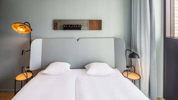 Una cama doble con ropa de cama blanca, dos mesitas de noche con lámparas, un estante en la pared con un letrero y una ventana con cortinas.