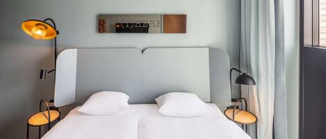 Un lit double avec du linge de lit blanc, deux tables de chevet avec des lampes, une étagère murale avec un panneau, et une fenêtre avec des rideaux.