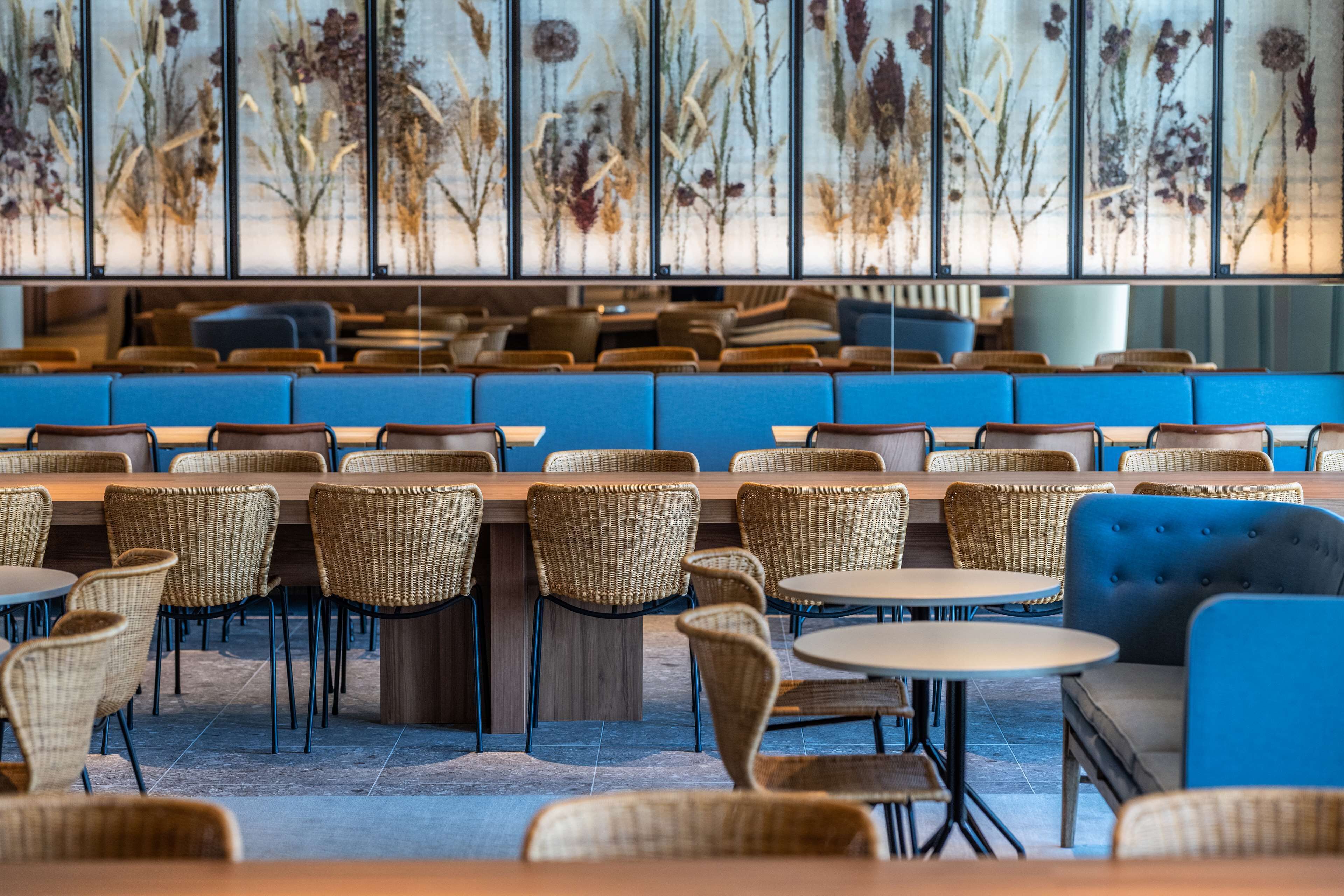 Een restaurant met rieten stoelen en tafels, blauwe zittingen met kussens, en een muur met bloemistische decoraties.
