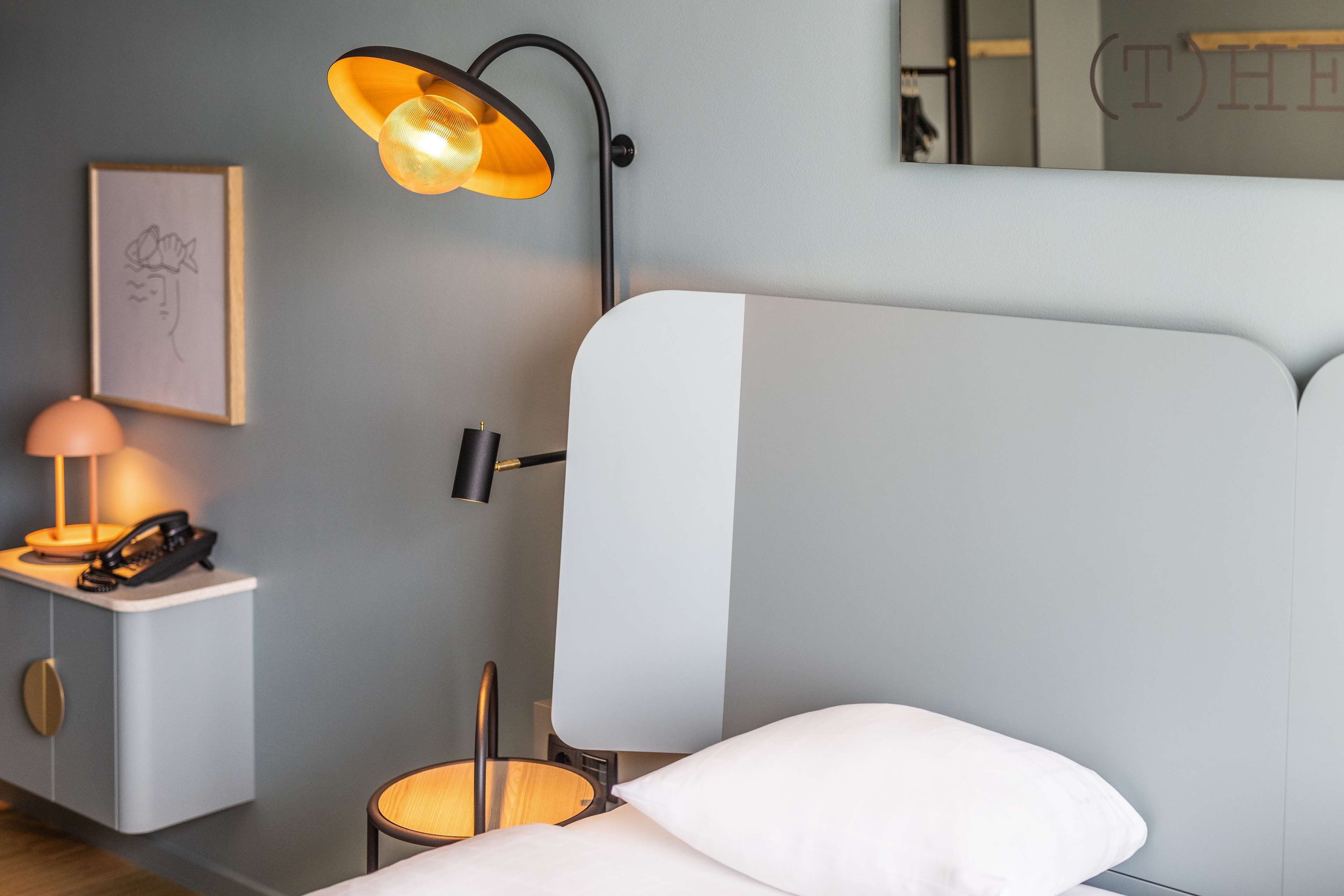 A modern hotel room with a bed, bedside lamp, nightstand, and a framed picture on the wall.
