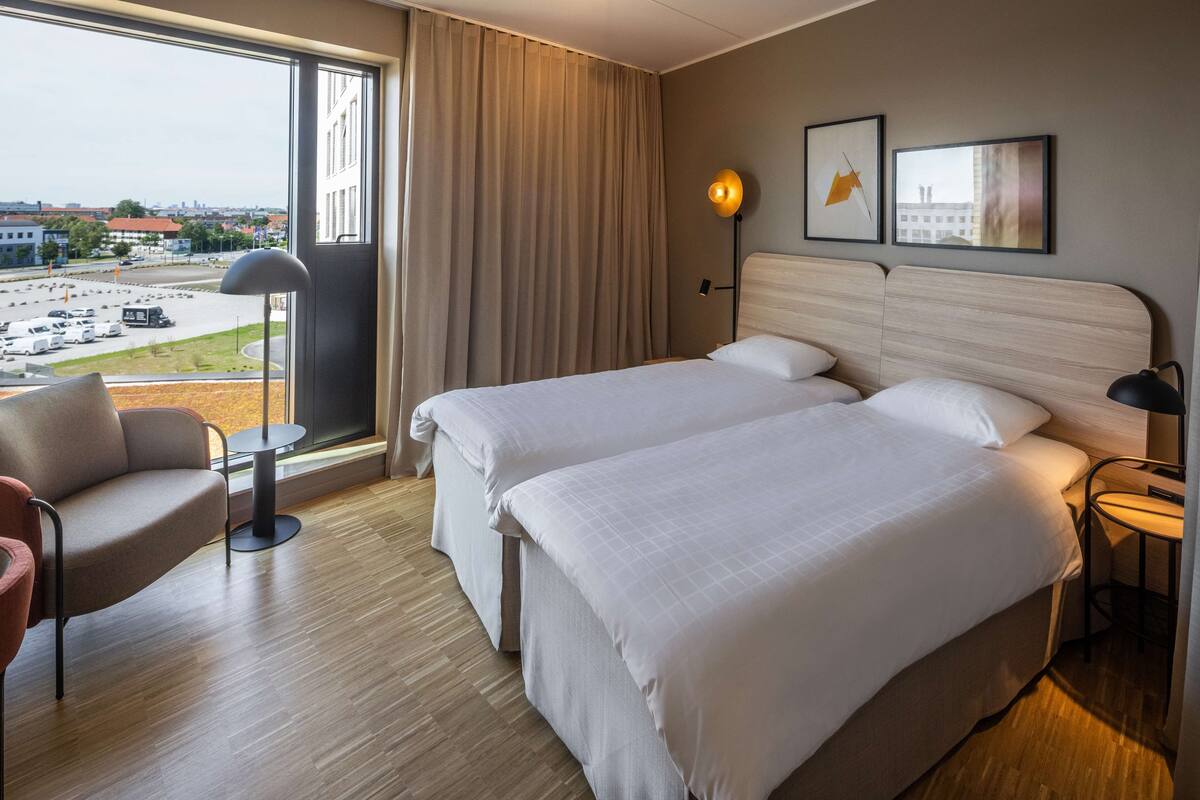 A hotel room with two beds, a view of a parking lot, a chair, and a lamp.