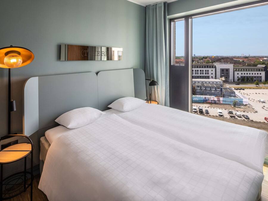 Una habitación de hotel con una cama grande, una mesita de noche, una lámpara, un espejo y vistas a la ciudad a través de un ventanal amplio.