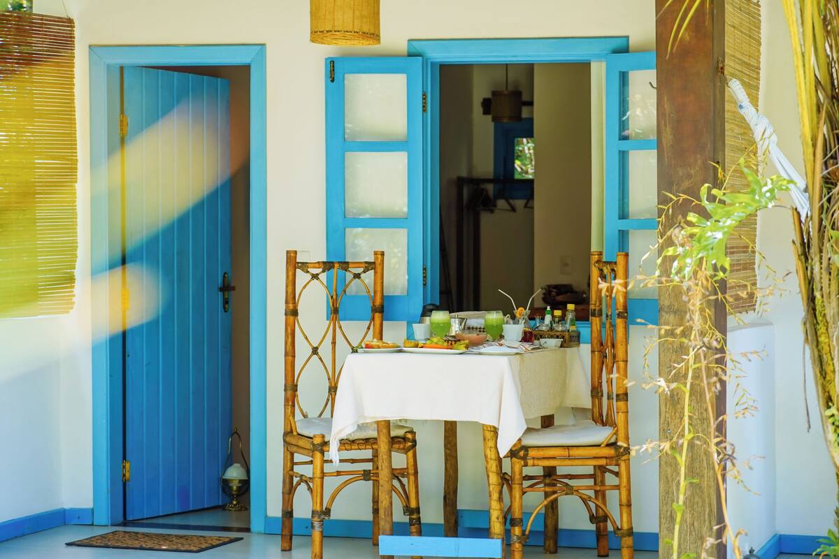 A dining area with a table set for two, surrounded by bamboo chairs, with a blue door and window in the background.