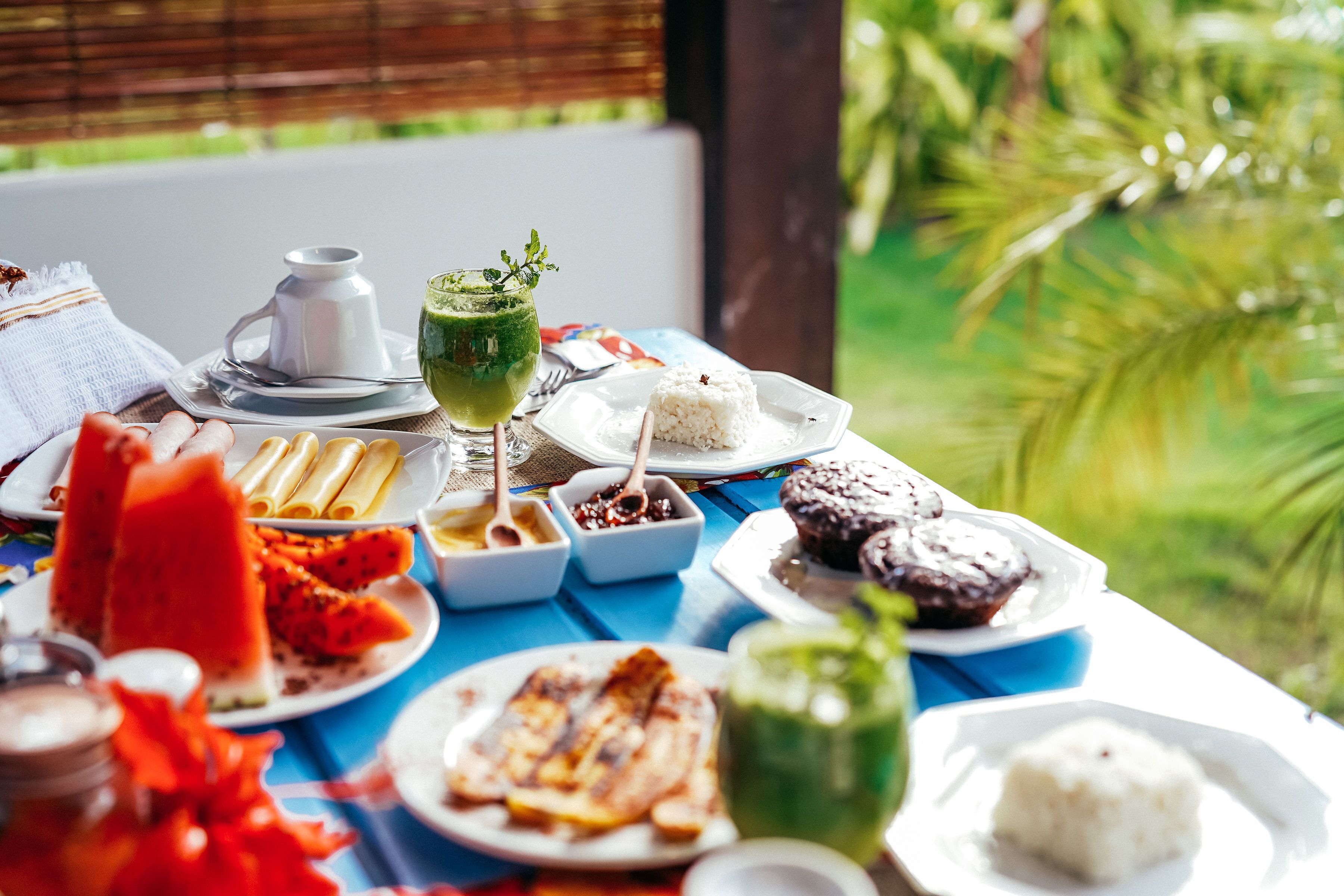 A table with various dishes including rice, grilled meat, and tropical fruits, set outdoors with a view of greenery.