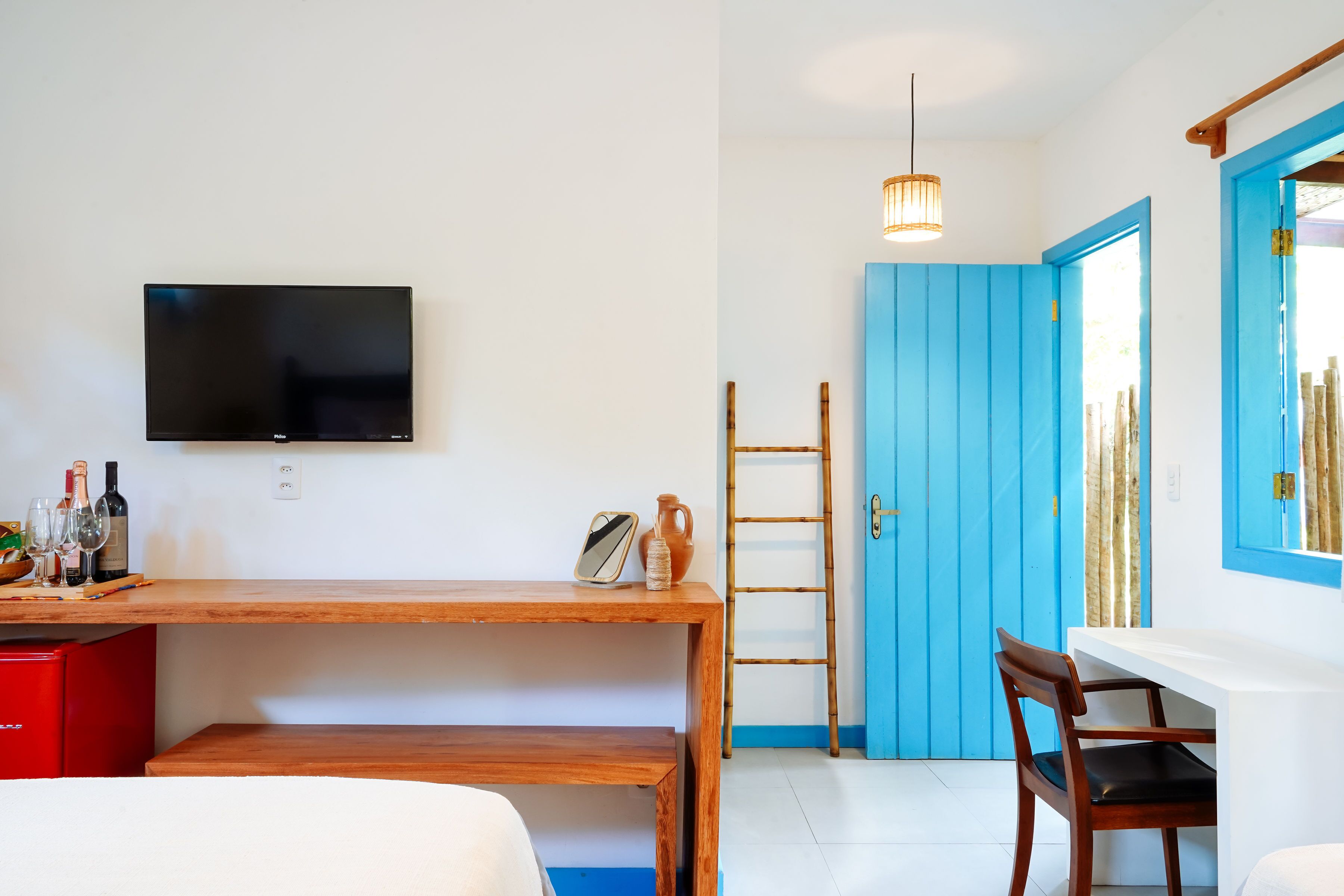 A room with a wooden desk, a television mounted on the wall, a red refrigerator, a blue door, and a wooden chair.
