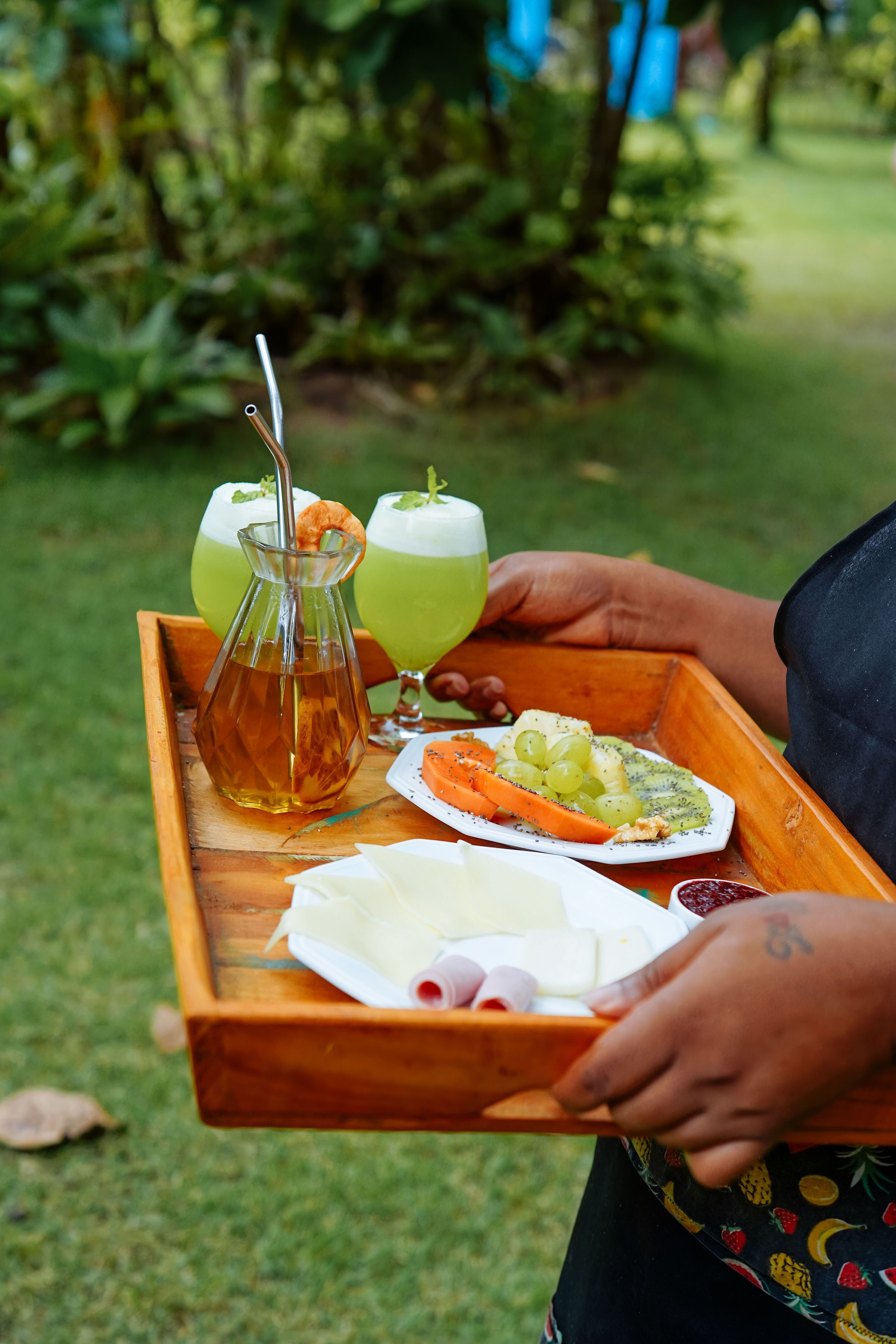 Una bandeja de madera con una jarra, dos vasos de bebida verde, un plato con uvas y queso, y un recipiente pequeño con salsa roja.