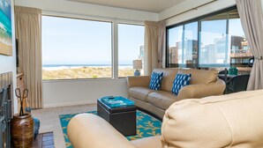 Ruime woonkamer met een groot raam dat uitzicht biedt op het strand en de gebouwen. Er staat een beige bank met gekleurde kussens, een klein poufje en een decoratieve vaas.