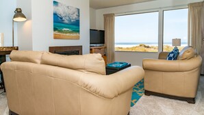 Een woonkamer met een beige bank, een kleinere fauteuil, een flatscreen-tv, een open haard en een groot raam met uitzicht op het strand.