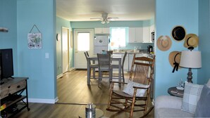 A living room with a television, a dining table with chairs, a rocking chair, and a lamp. There is a kitchen in the background with cabinets and a refrigerator.