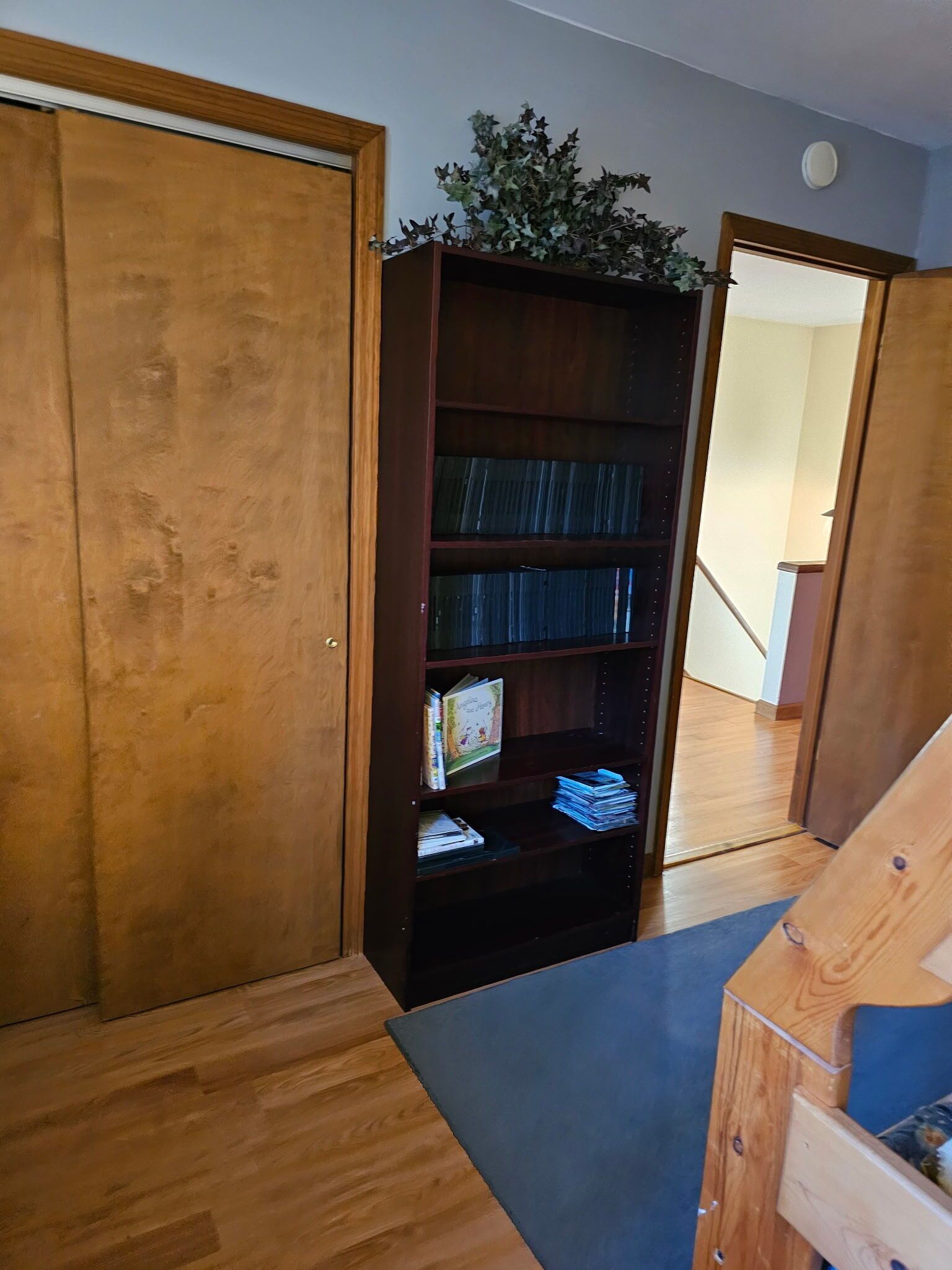 A room with a wooden floor, a bookshelf with books and decorative items, and a door leading to another room.