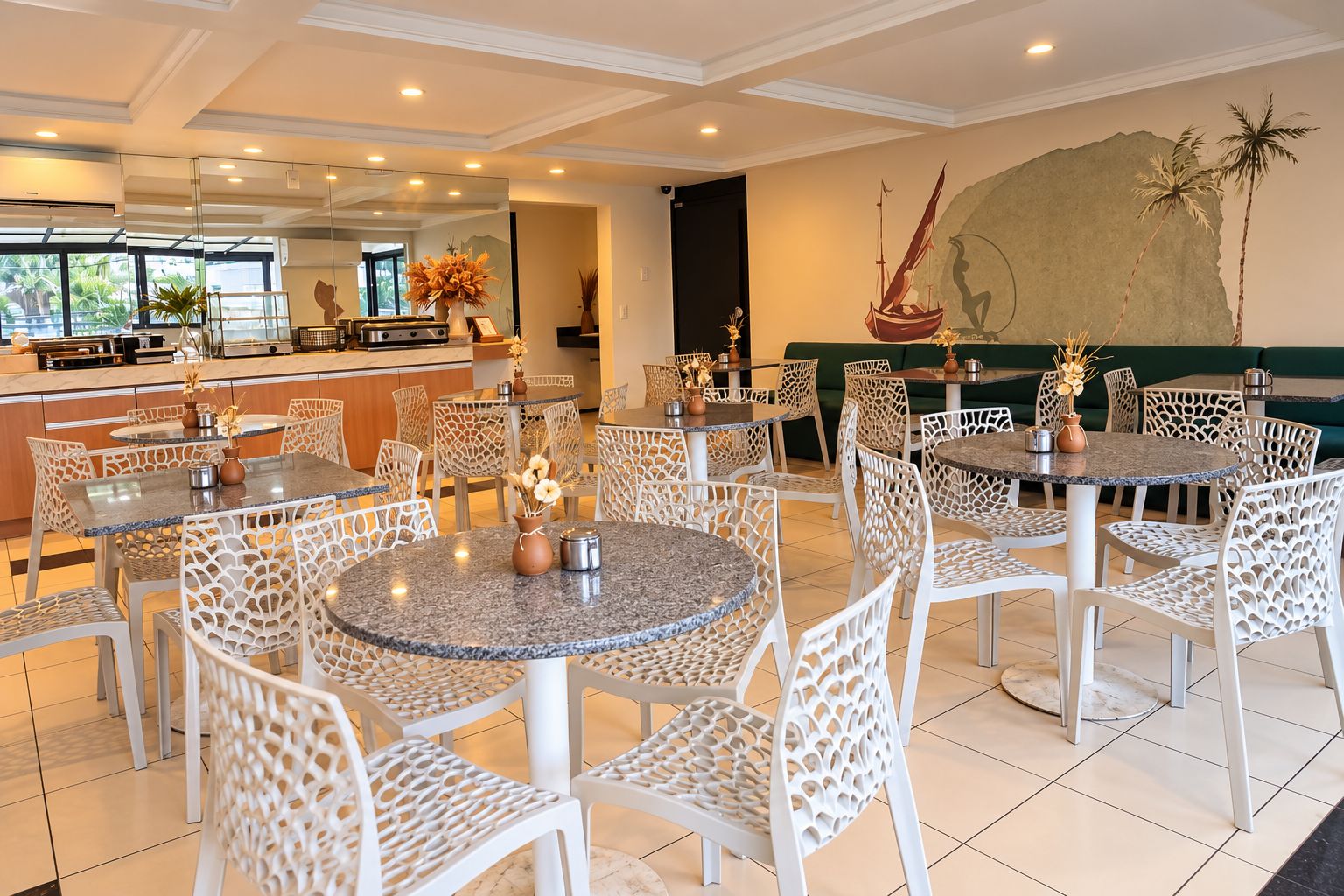 A hotel dining area with round tables and chairs, a kitchen in the background, and a mural on the wall.