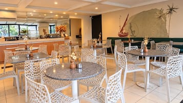 A hotel dining area with round tables and chairs, a kitchen in the background, and a mural on the wall.