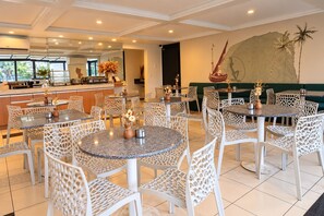 Comedor de hotel con mesas y sillas redondas, una cocina al fondo y un mural en la pared.
