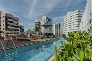 Uma piscina com espreguiçadeiras e uma mesa, cercada por edifícios modernos e um céu claro.