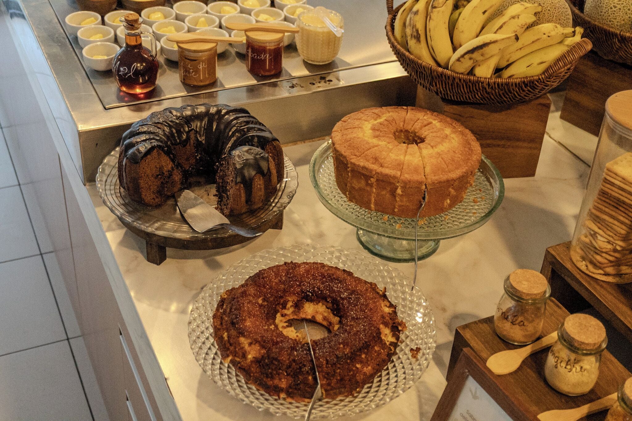 A bakery display with various cakes and pastries, including a chocolate bundt cake, a vanilla bundt cake, and a fruit-topped cake.