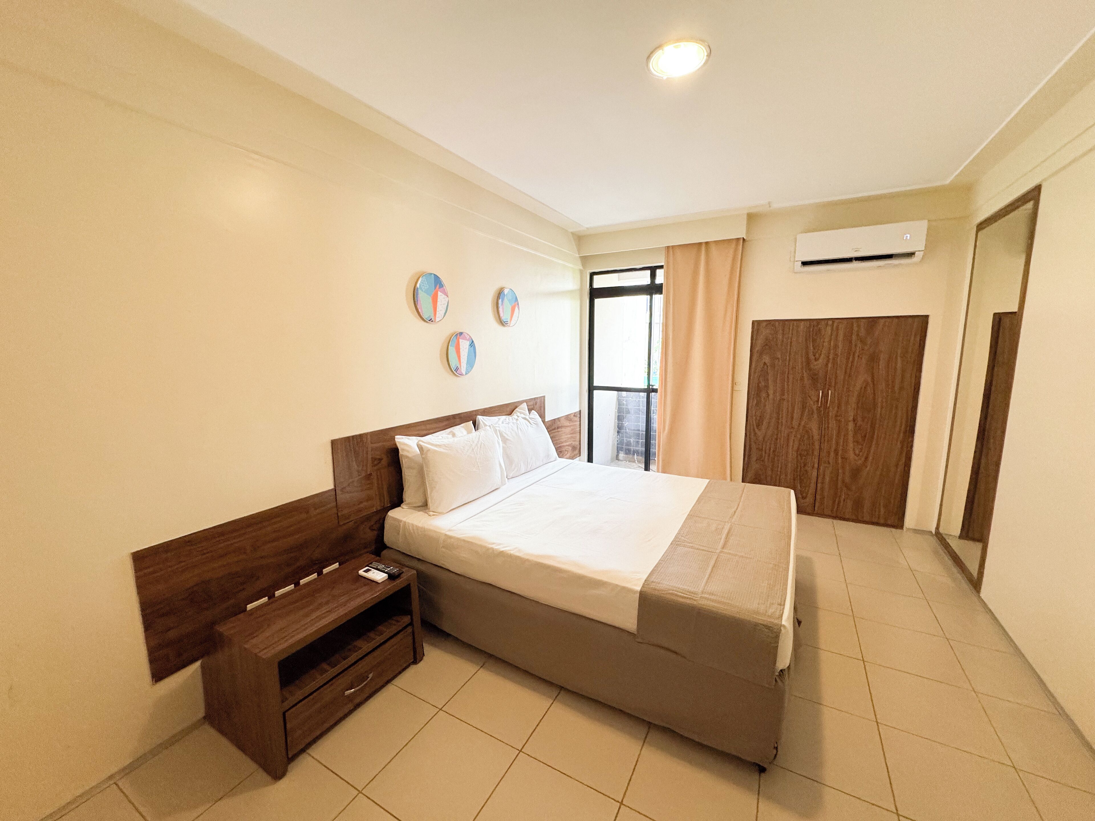 A hotel room with a bed, a wooden nightstand, a wardrobe, and an air conditioning unit.