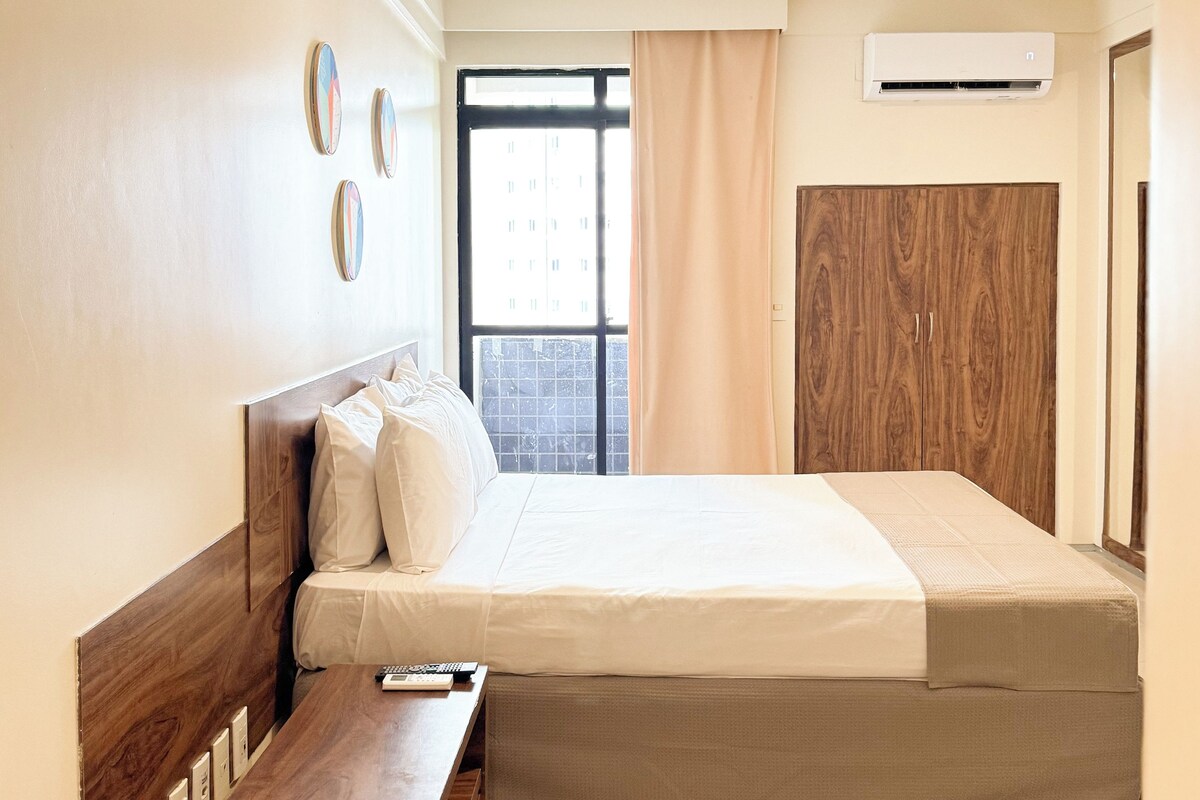 Une chambre d’hôtel avec une tête de lit en bois, un lit avec une literie blanche, une table de chevet, une fenêtre avec des rideaux et une armoire en bois.