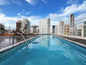 Une piscine sur le toit, accessible par un escalier en métal, entourée de bâtiments modernes et surplombant un ciel dégagé.