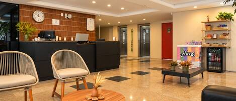 Un hall de hotel con una recepción, dos sillones, una mesa de centro y un refrigerador.