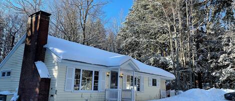 A single-story house with a brick chimney, a porch, and a snow-covered roof.