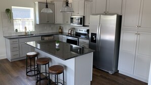 Una cocina moderna con armarios blancos, electrodomésticos de acero inoxidable, una isla central con taburetes, iluminación colgante y una ventana con vistas.