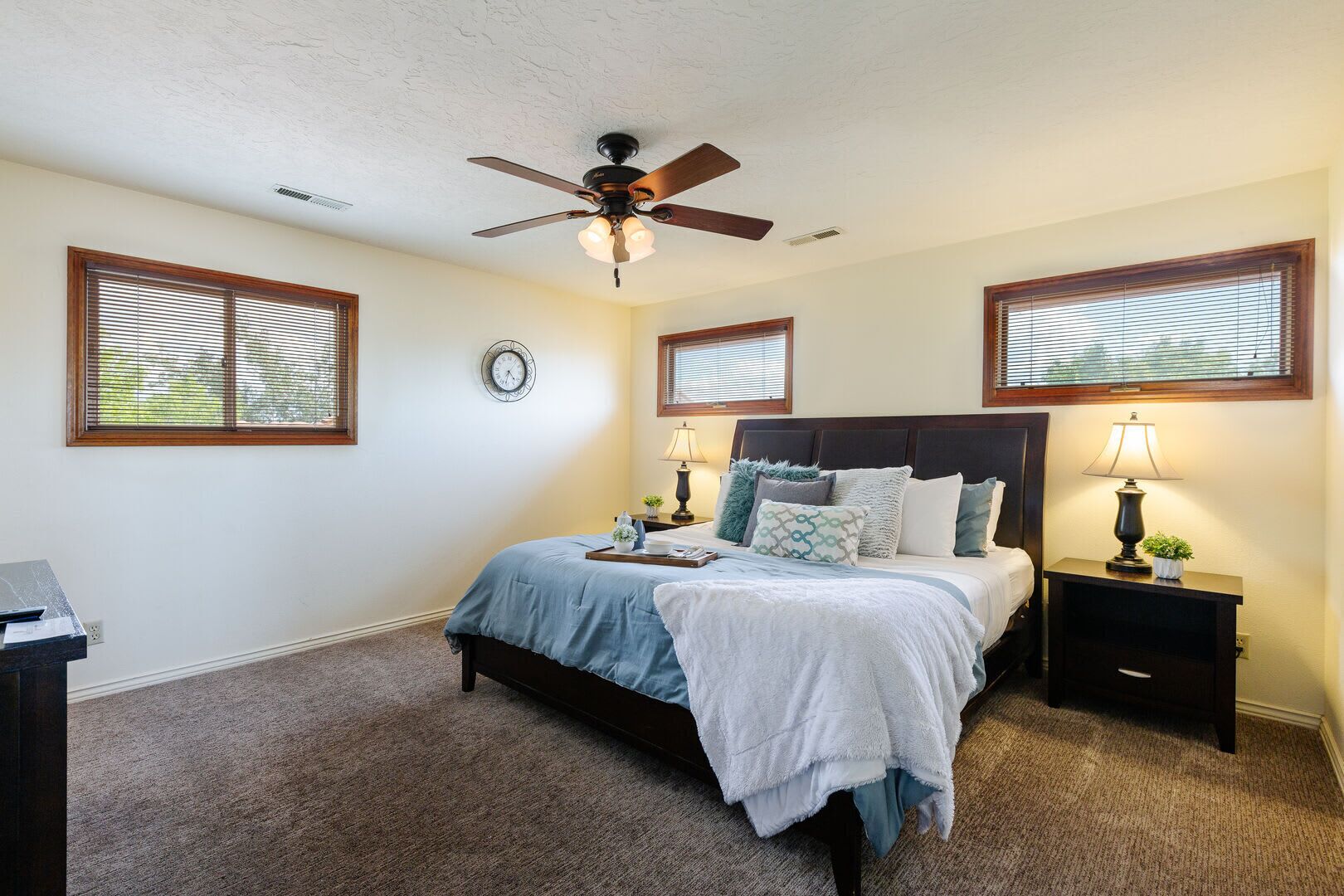 A bedroom with a bed, two bedside tables, a ceiling fan, and three windows with blinds.