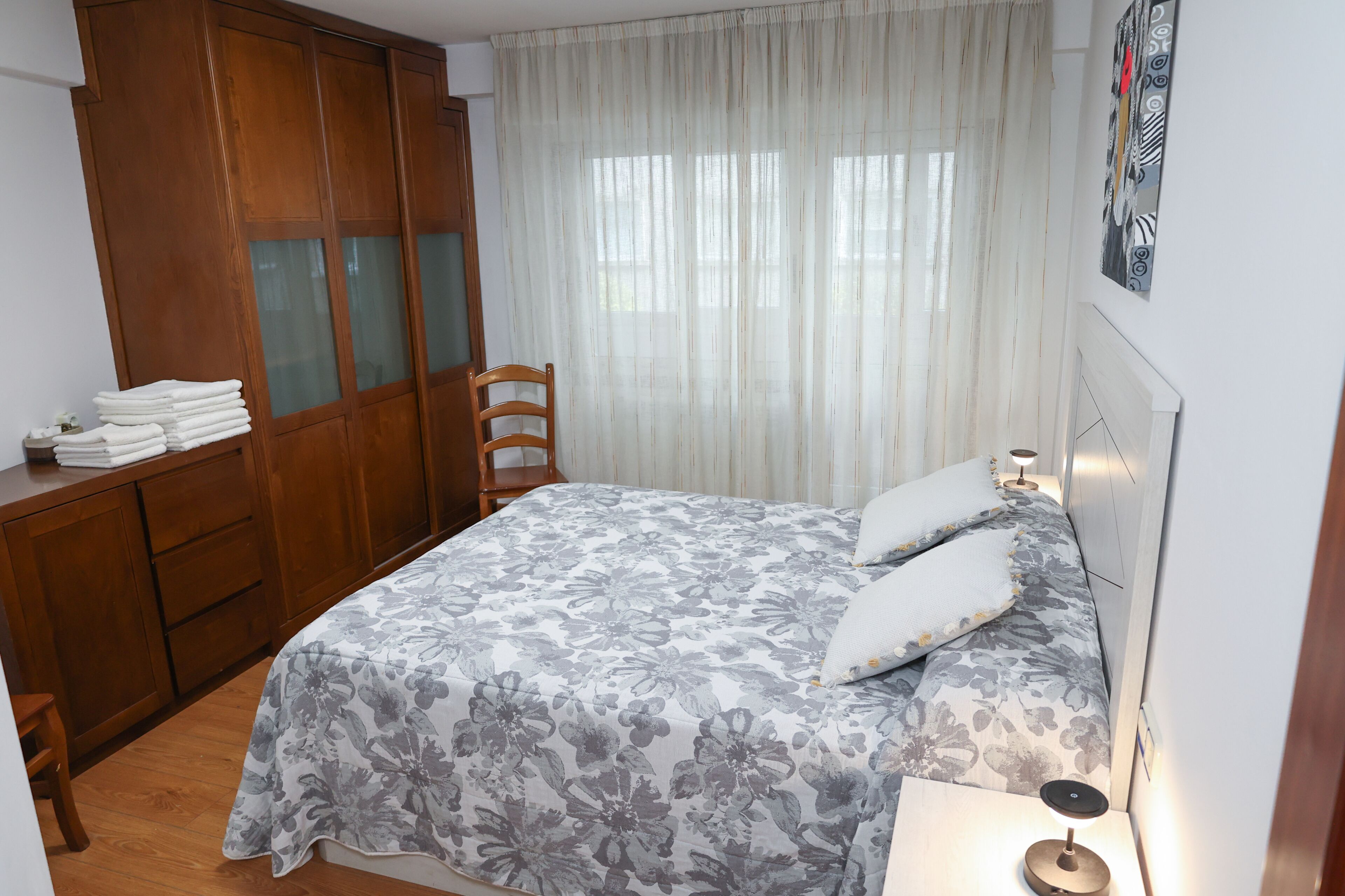 Un dormitorio con cama, armario de madera, silla y mesita de noche con lámpara.