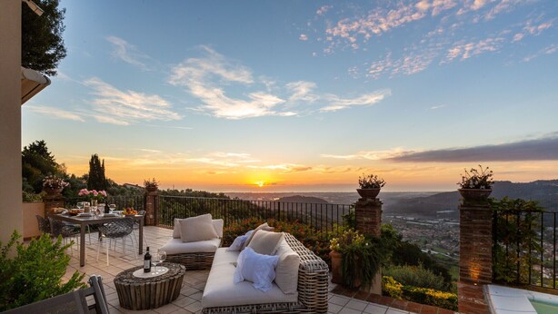 Une terrasse extérieure aménagée avec des meubles en osier, une table à manger et une vue sur le coucher de soleil au-dessus d’un paysage urbain.